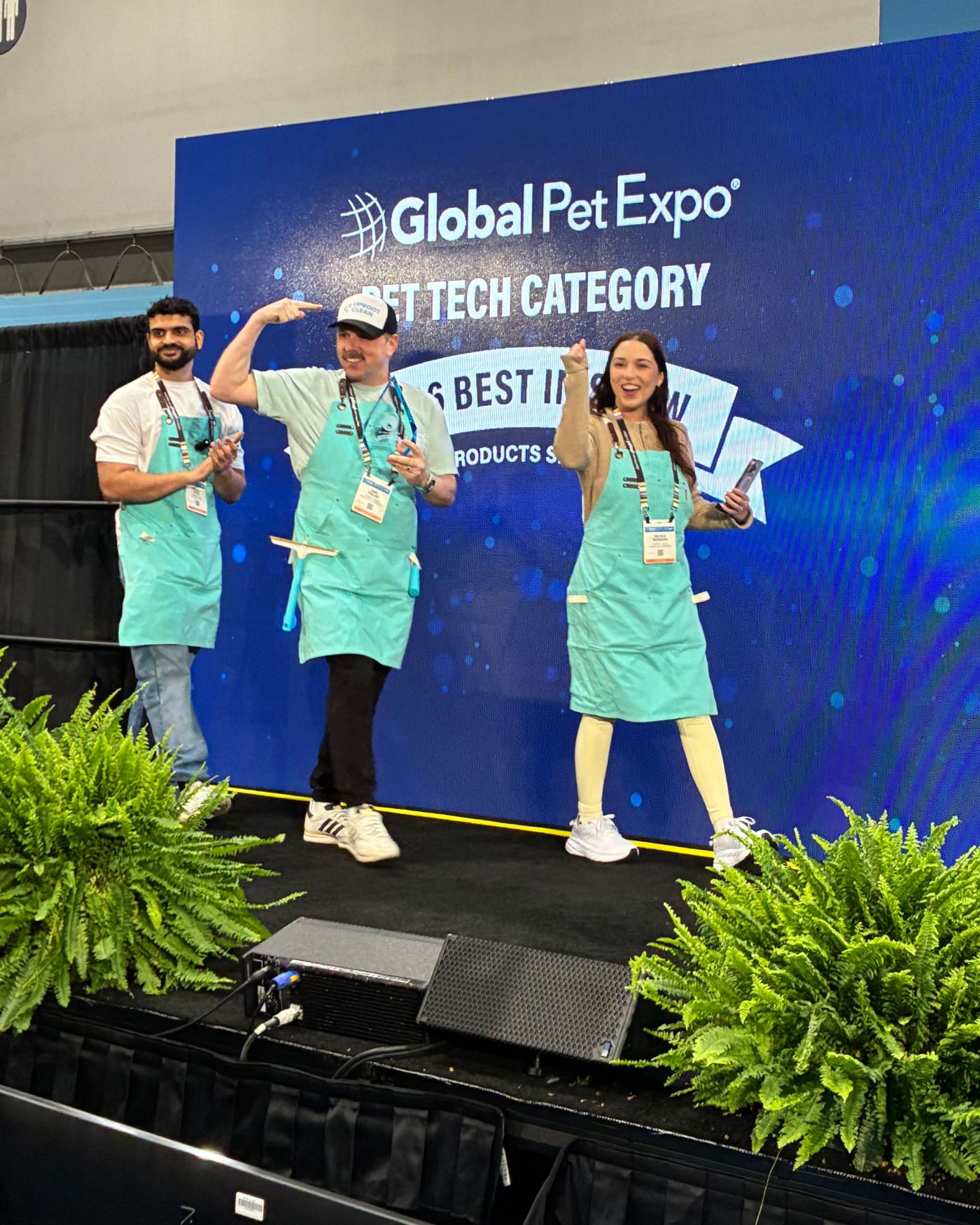 Three Uproot Clean team members in teal aprons celebrating on stage under the Global Pet Expo Pet Tech Category banner